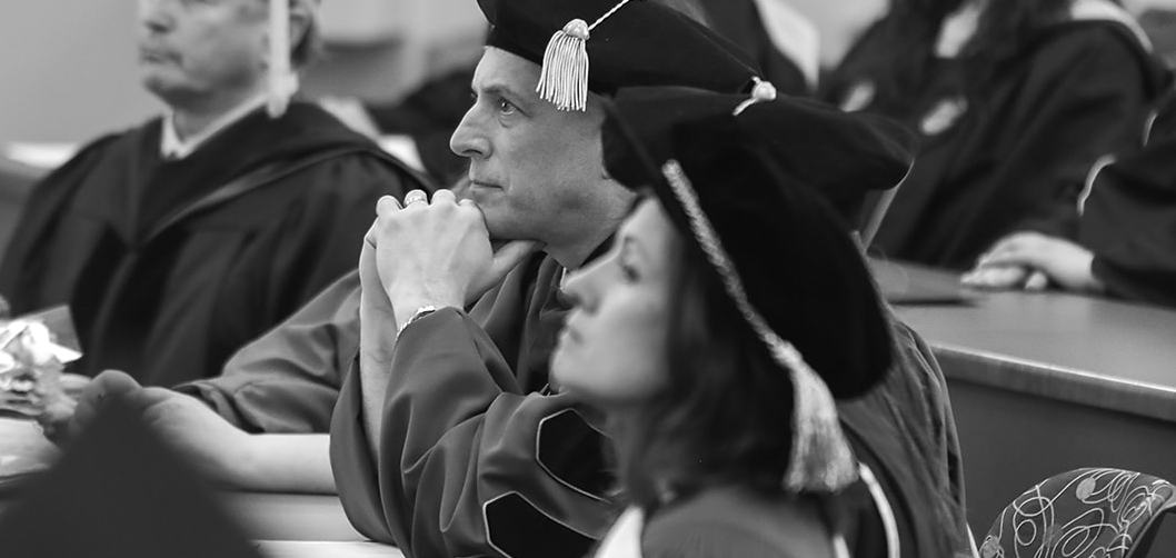 A black and white image of people in graduation regalia seated in a classroom and looking forward
